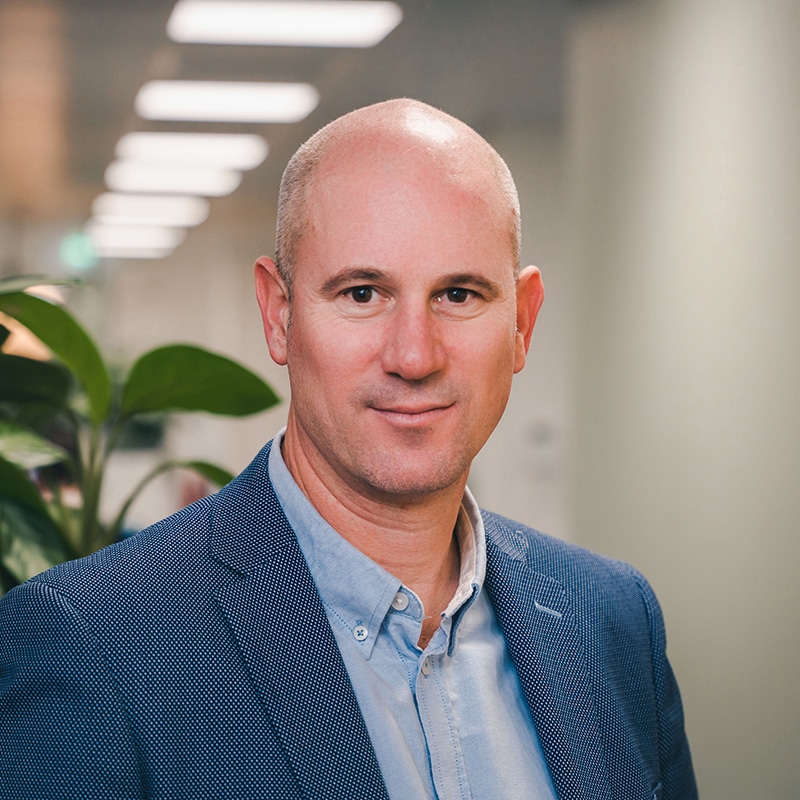 Headshot of Byron Tarboton wearing a pale blue shirt under a dark blue blazer in front of a white wall and green plants.