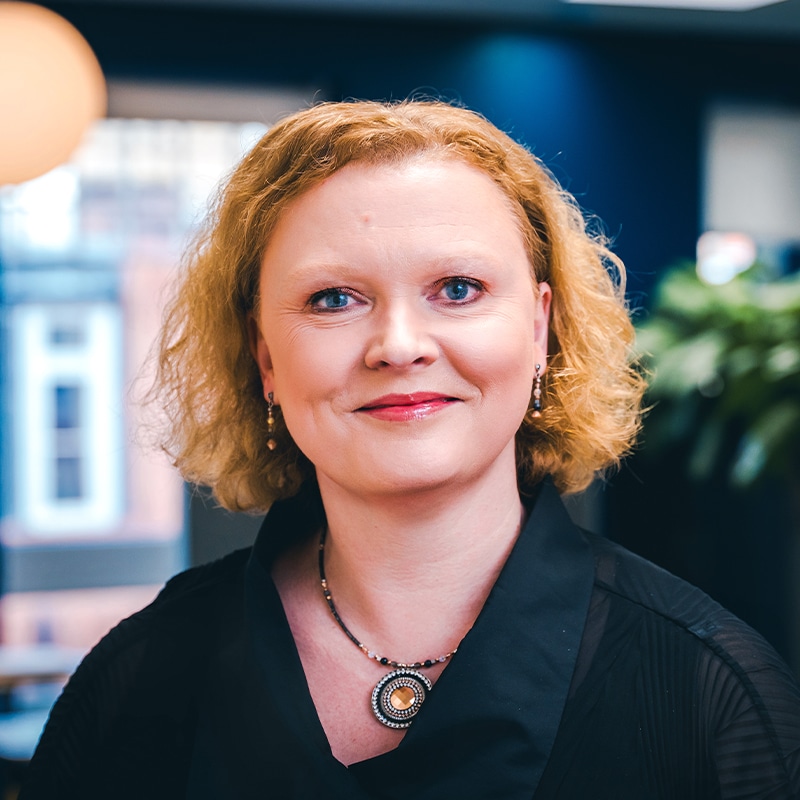 Headshot of Emma-Claire Kavanagh wearing a black shirt with a black and orange necklace in front of a blue wall and green plants.