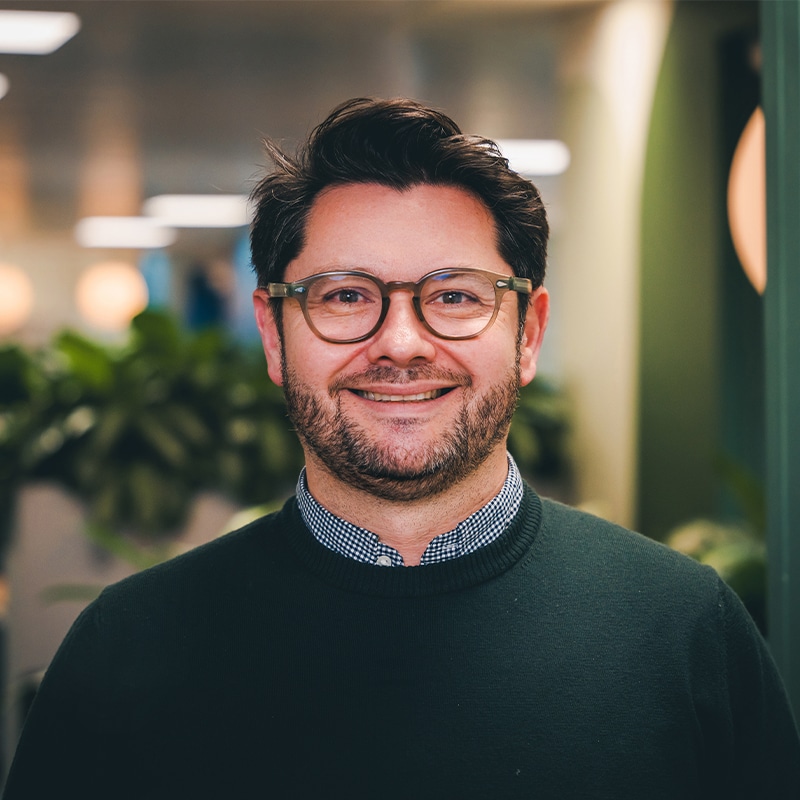 Headshot of Gordon Whyte, CEO at BIE, wearing a green jumper with a black and white shirt in front of a green wall and some green plants in the background.