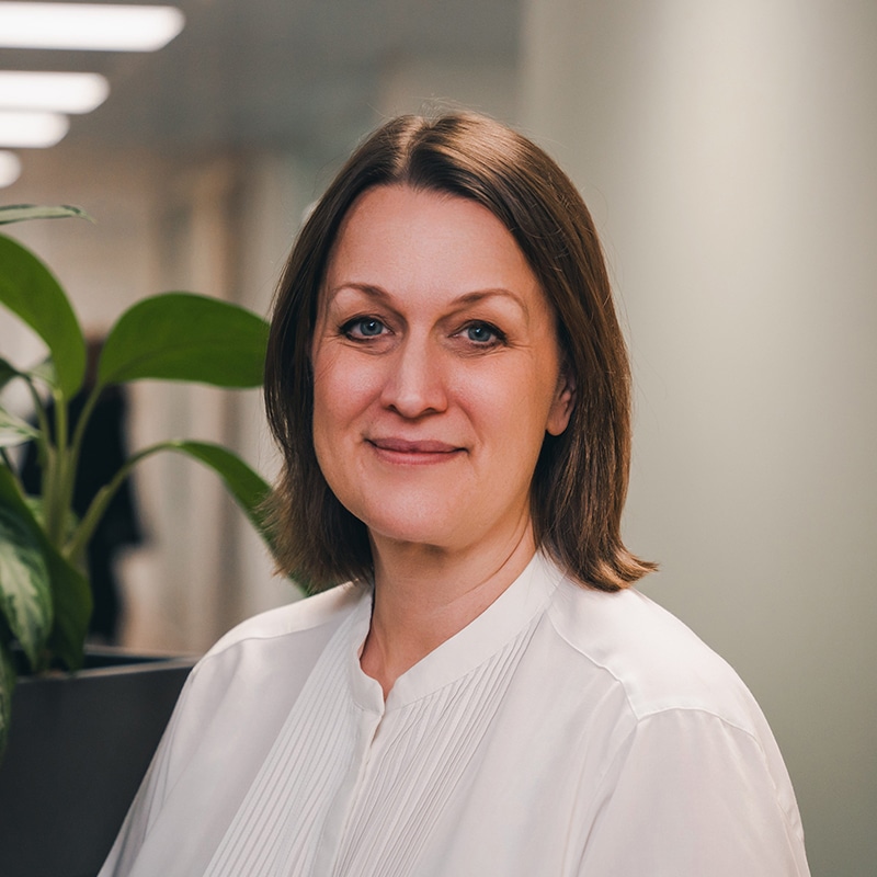 Headshot of Kate Mathias, a NED at BIE, wearing a white shirt in front of a white wall and some green plants in the BIE office.