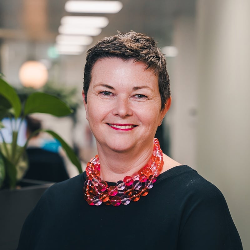 Headshot of Lisa Vigurs, Chief of Staff at BIE, wearing a black top with a pink and orange necklace and pink lipstick, in front of some plants and a white background.