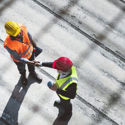 Two construction workers wearing hard hats shaking hands. One is wearing a yellow hat and orange hi-vis vest, the other is wearing a red hat and a yellow hi-vis vest.