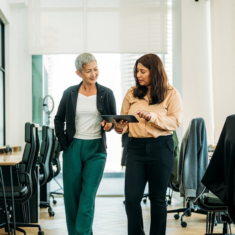 Two female colleagues smiling and having a conversation in a corridor over an iPad. One is an older asian woman with short grey hair wearing a dark blazer and a white top. The other is a south asian woman wearing a light blouse.