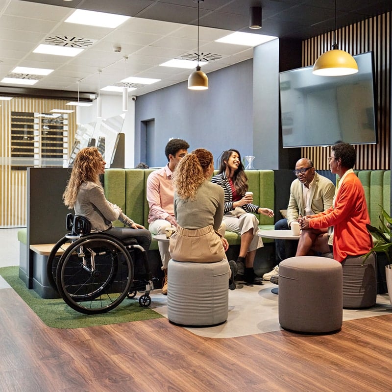 Group of colleagues of mixed ethnicity and ages sat on benches and stools having a conversation and smiling. One female colleage with blonde hair is in a wheelchair.