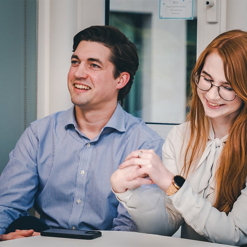 Alex Hyde, from BIE, wearing a blue shirt and smiling, sitting next to Maddie McCoubrey, from BIE, wearing glasses and a white blouse looking down.