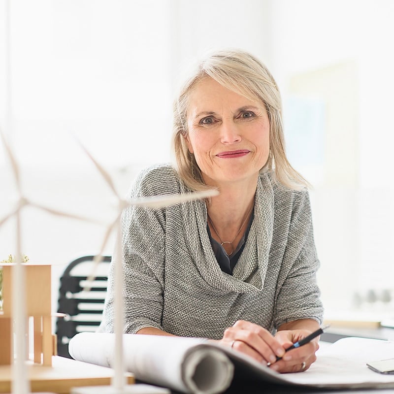 An older business woman, sitting down and smiling into the camera. She has blonde grey hair and is wearing a grey loose fitting jumper.