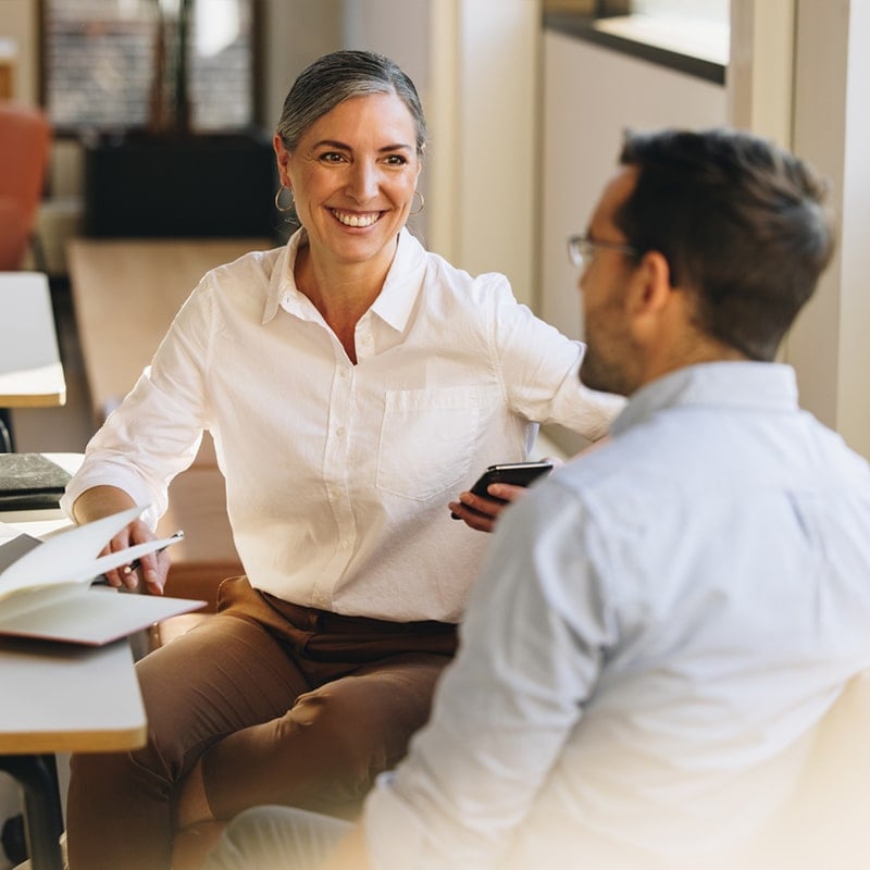 A business woman smiling at a male colleague in a conversation with him. She is middle aged, with slightly grey hair, holding a mobile phone and wearing a white shirt. He has his back to the camera and is wearing a blue shirt.