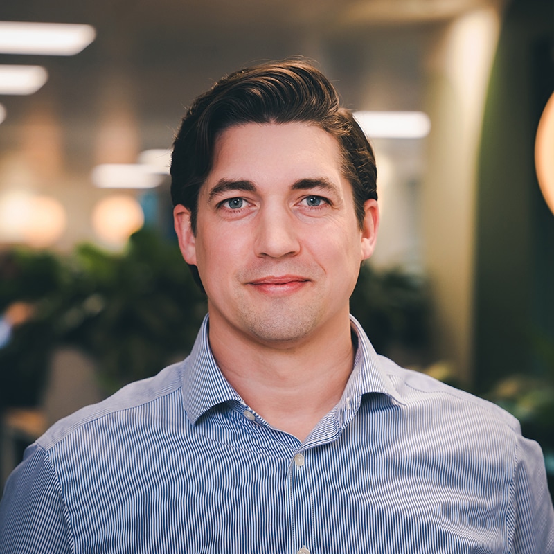 Headshot of Alex Smith, wearing a blue and white pinstripe shirt in front of a green wall and green plants.