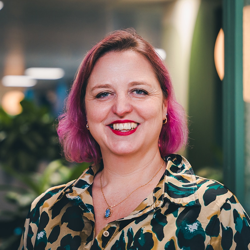 Headshot of Ann-Maree, with pink hair and lipstick, wearing a gold, green and black leopard print top and a blue and gold necklace, standing in front of a green wall and green plants.