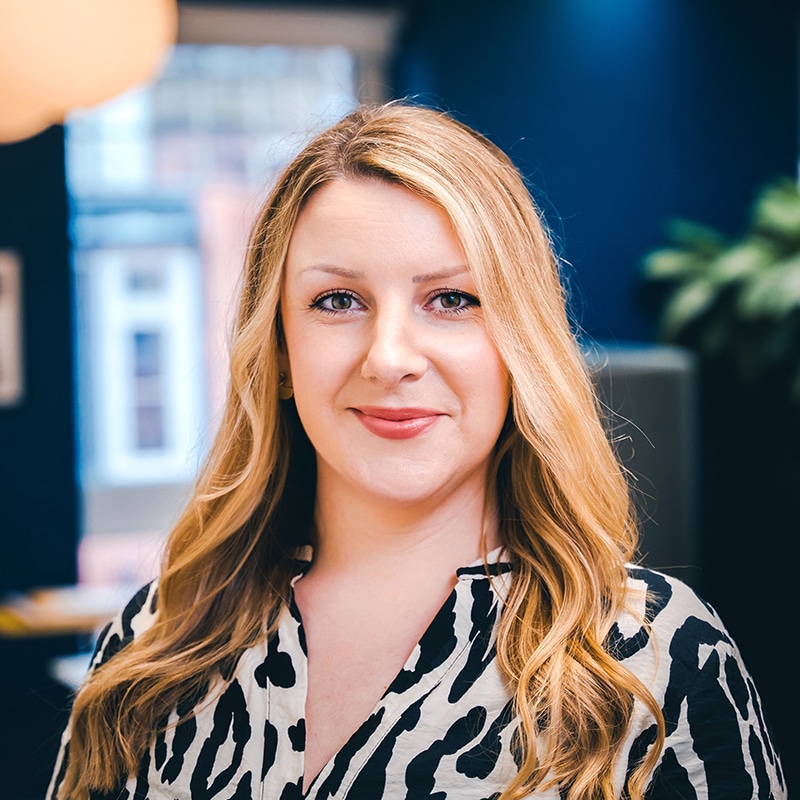 Headshot of Anna Davies wearing a black and white leopard print shirt in front of green plants and a blue wall.