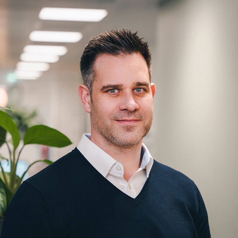 Headshot of Ben Bond wearing a white shirt underneath a blue V-neck jumper in front of a white wall and green plants.