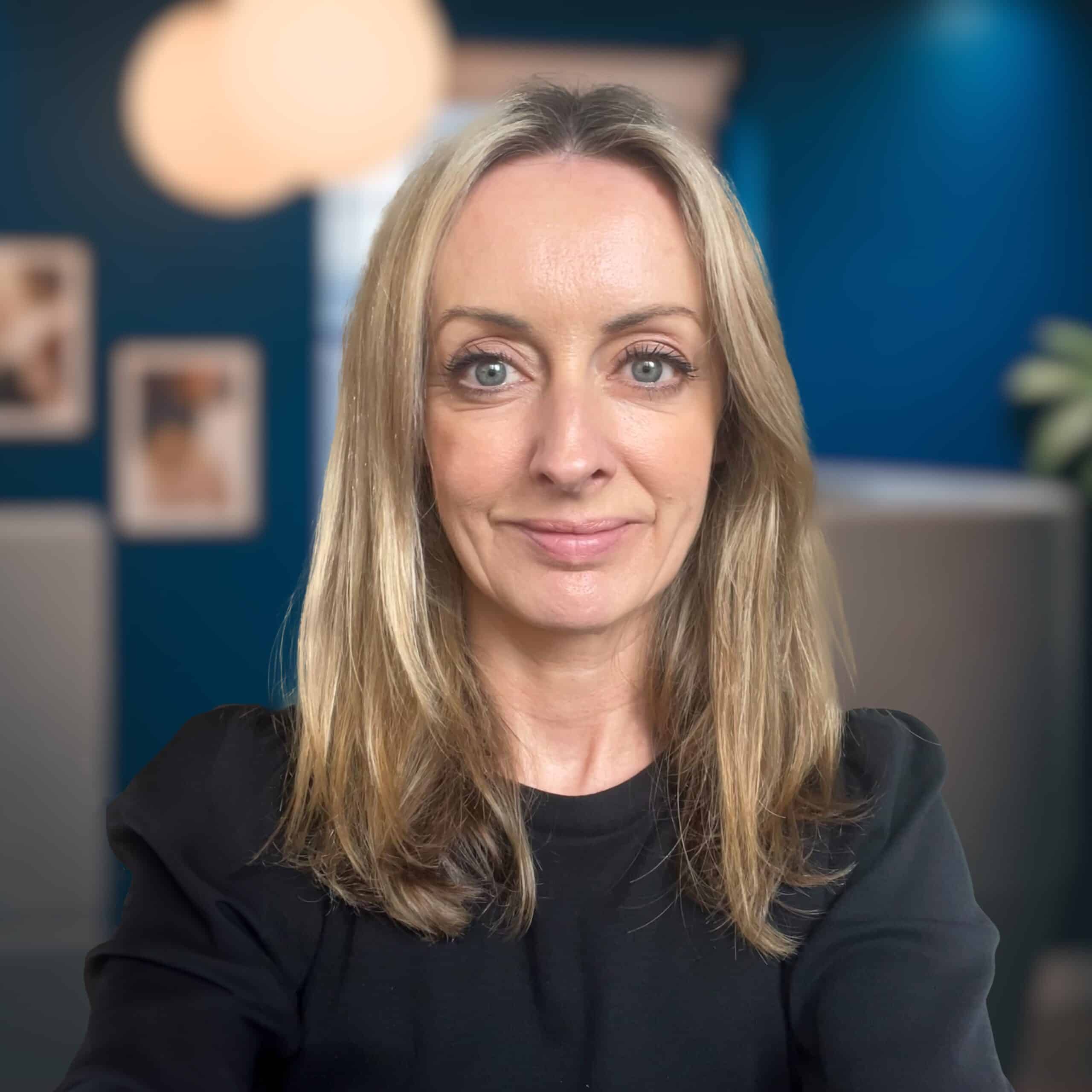 Headshot of Claire Paramo, Managing Director at BIE, wearing a black sweater, in front of a blue wall and two grey seats in the BIE office.