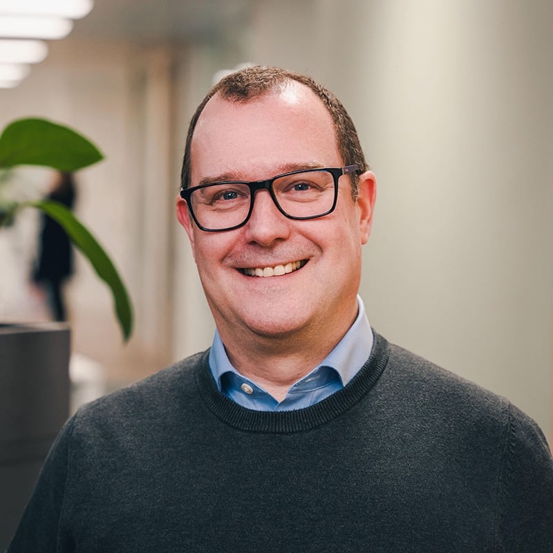 Headshot of Cyr Cornberg wearing a blue shirt underneath a grey jumper with black glasses, in front of a white wall and green plants.