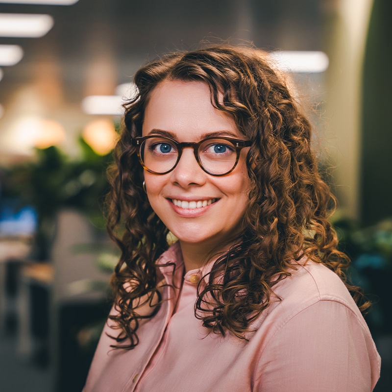 Headshot of Daisy Goodey, wearing a light pink shirt and brown glasses with curly hair in font of a green wall and green plants.