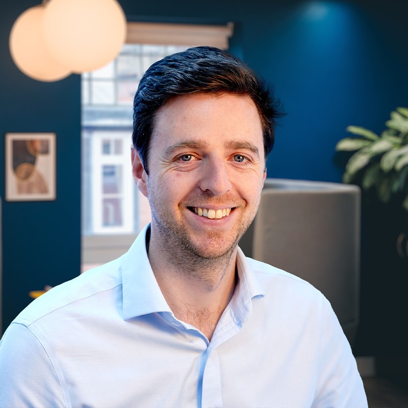 Headshot of Dan Middleton wearing a light blue shirt in front a grey seating area with blue walls and green plants