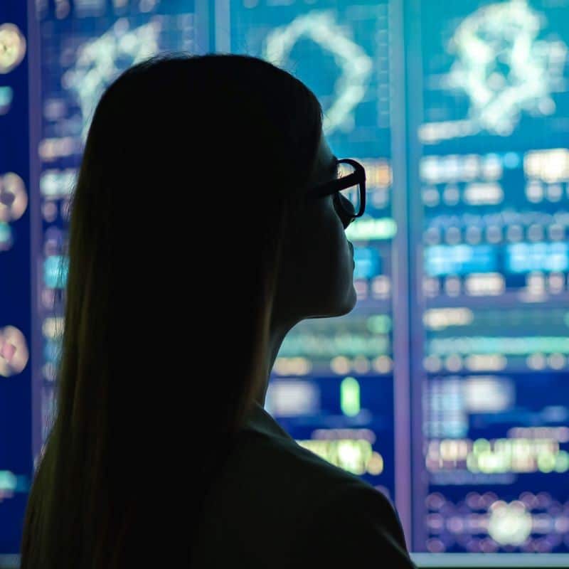 Woman wearing glasses standing in front of a large screen with lots of data on it.