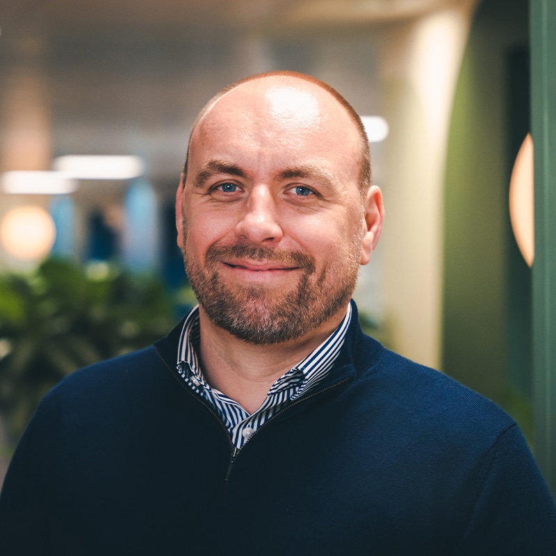 Headshot of Gareth Paul wearing a blue and white pinstripe shirt underneath a blue quarter-zip jumper in front of a green wall and green plants.