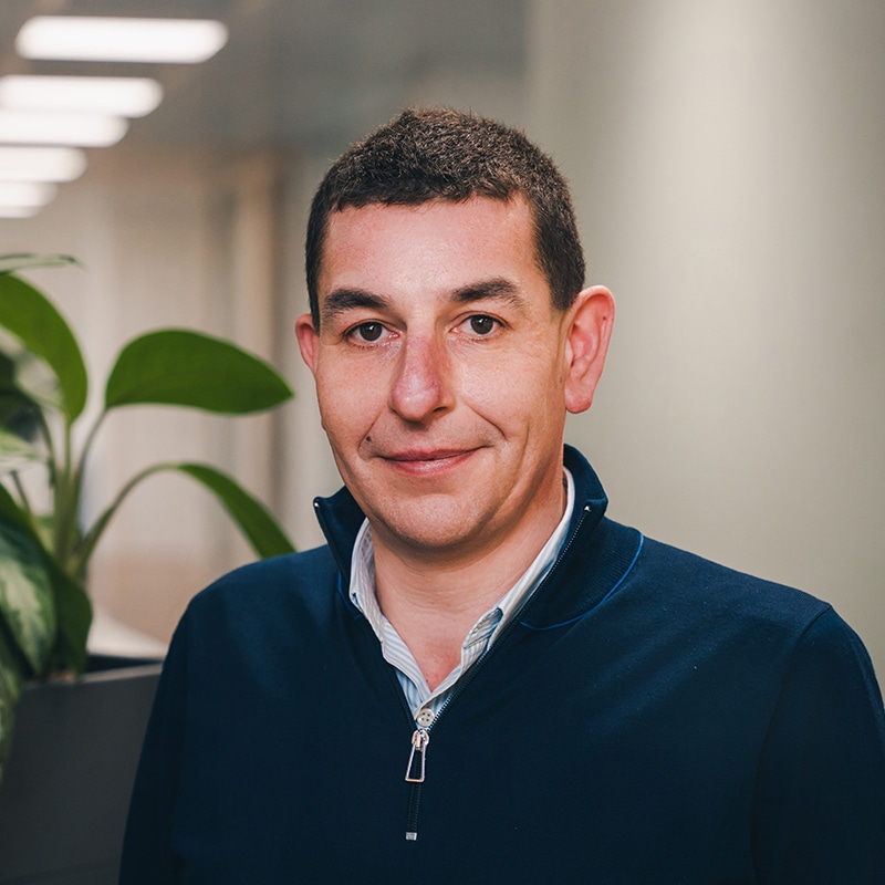 Headshot of James Gherardi wearing a blue and white pinstripe shirt underneath a blue quarter-zip jumper in front of a white wall and green plants.