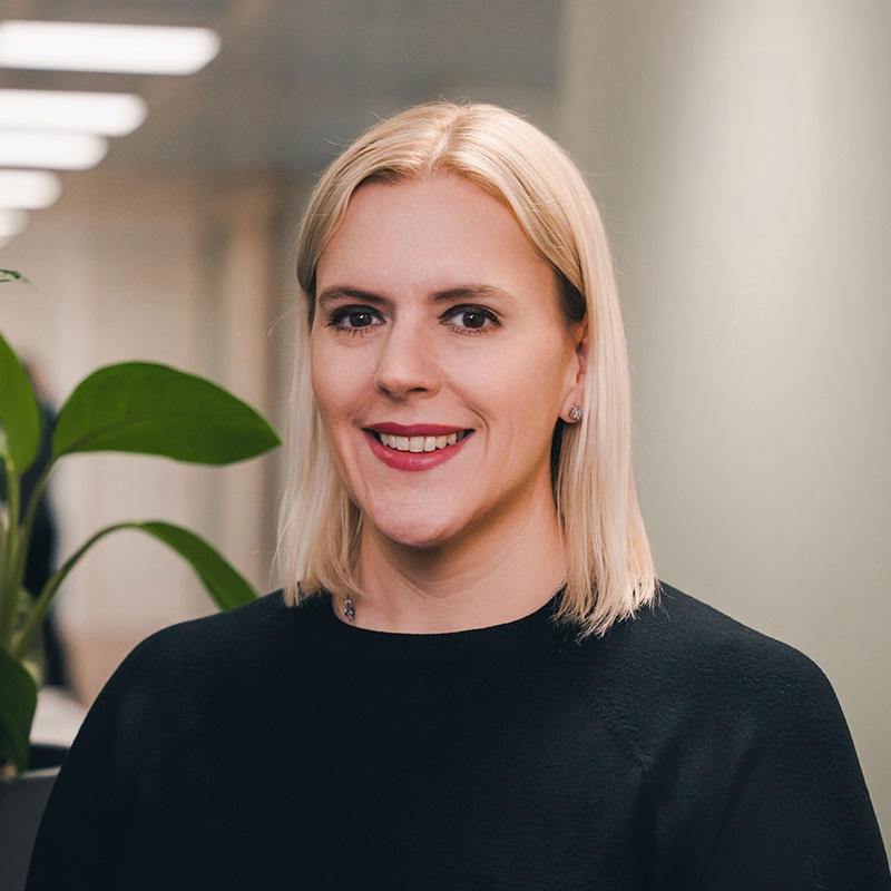 Headshot of Jenny Gardiner wearing a black sweater in front of a white wall and green plants.