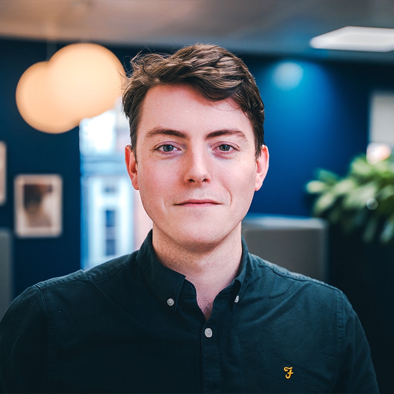 Headshot of Kai Fogarty wearing a blue shirt in front of a blue wall and green plants.