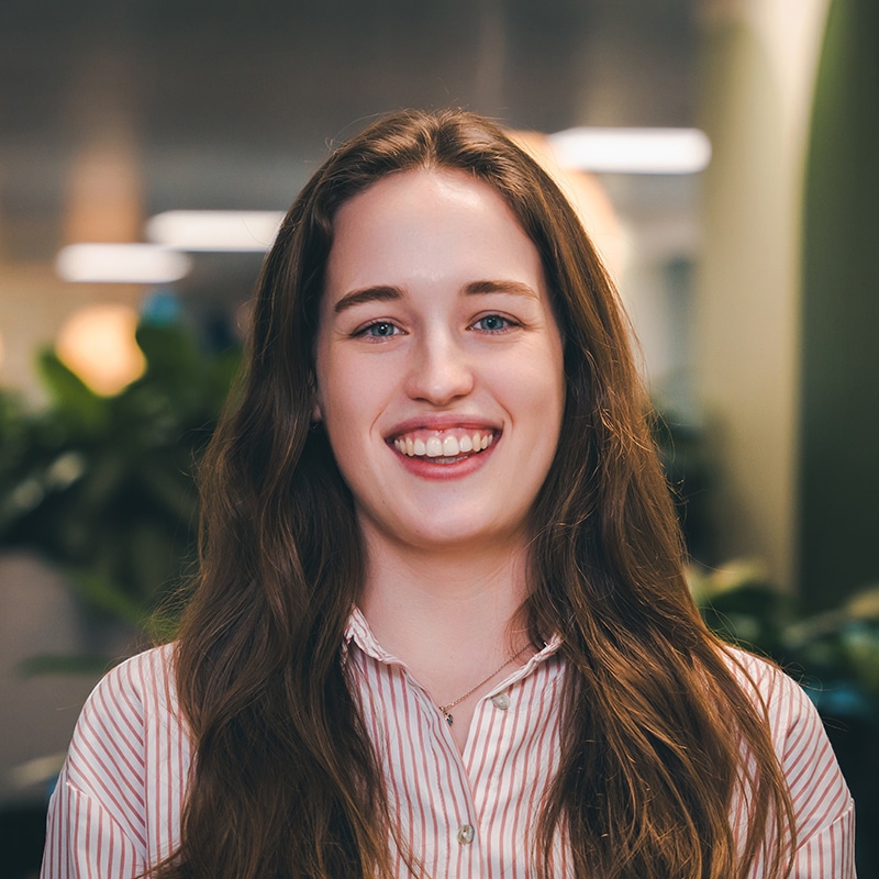 Headshot of Kate Mathias wearing a pink and white pinstripe shirt in front of a green wall and green plants.