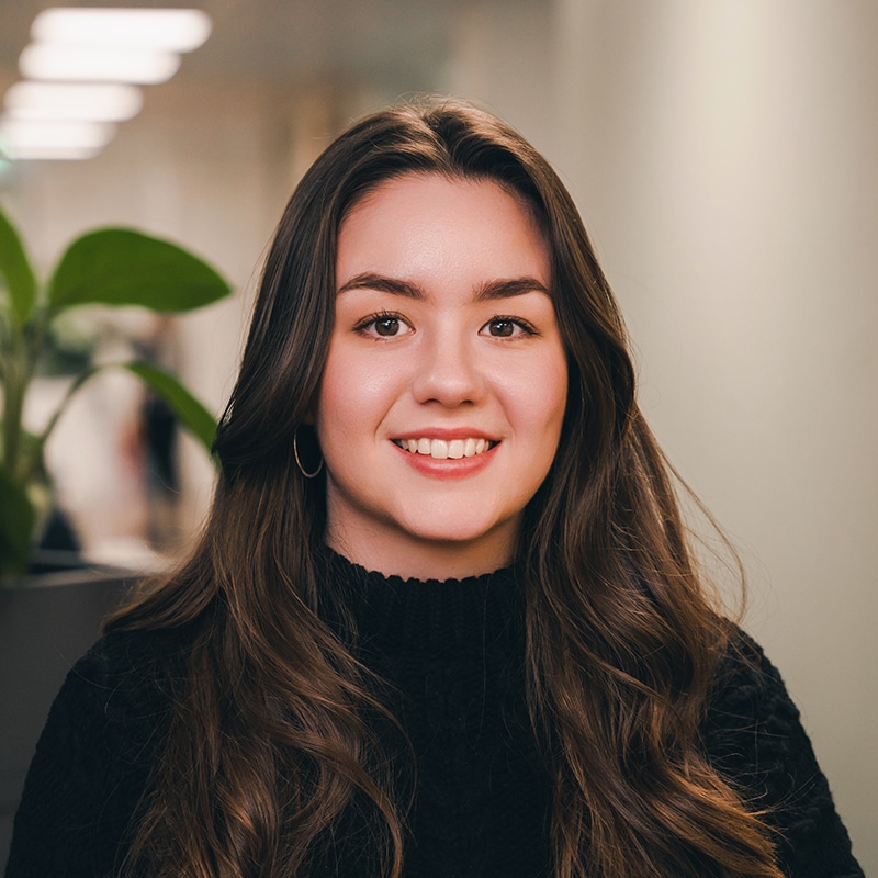 Headshot of Lia Trunchion wearing a black sweater with curly hair in front of a white wall and green plants.