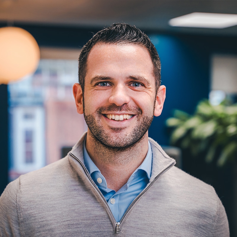 Headshot of Luke Moriarty, wearing a blue shirt underneath a grey quarter-zip jumper in front of a blue wall and green plants.