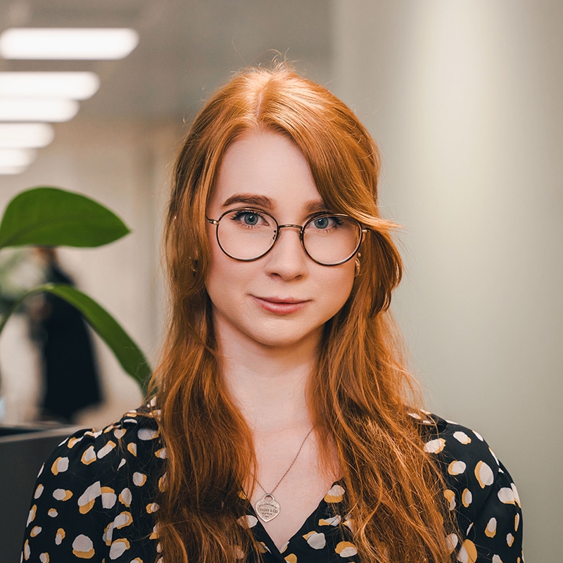 Headshot of Maddie McCoubrey, wearing a black top with white and yellow spots, and a gold necklace and glasses, in front of a white wall and green plants.