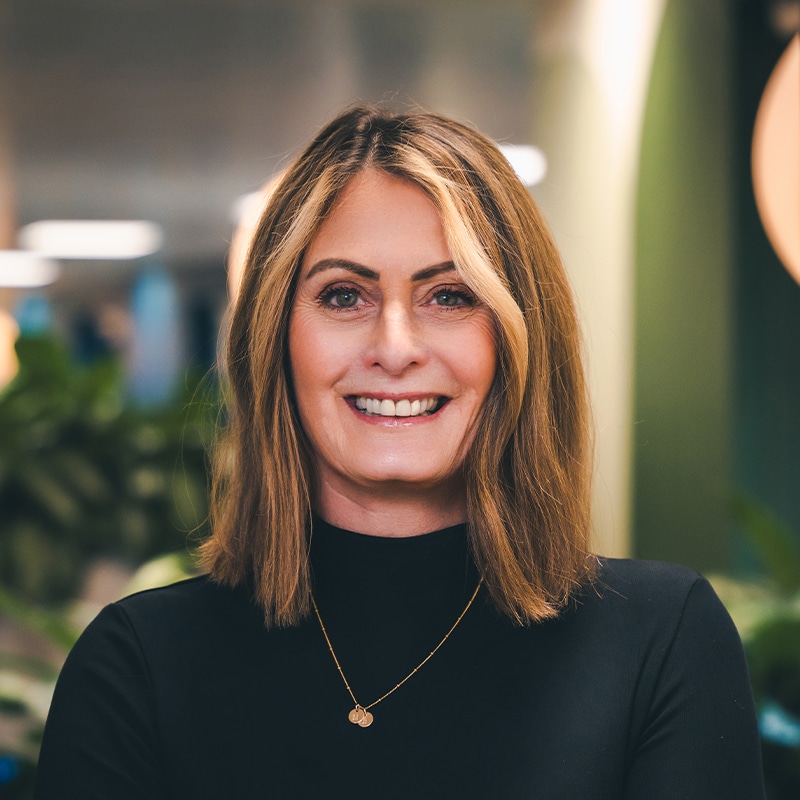 Headshot of Mandy Scarff wearing a lack high-neck top with a gold necklace in front of a green wall and green plants.