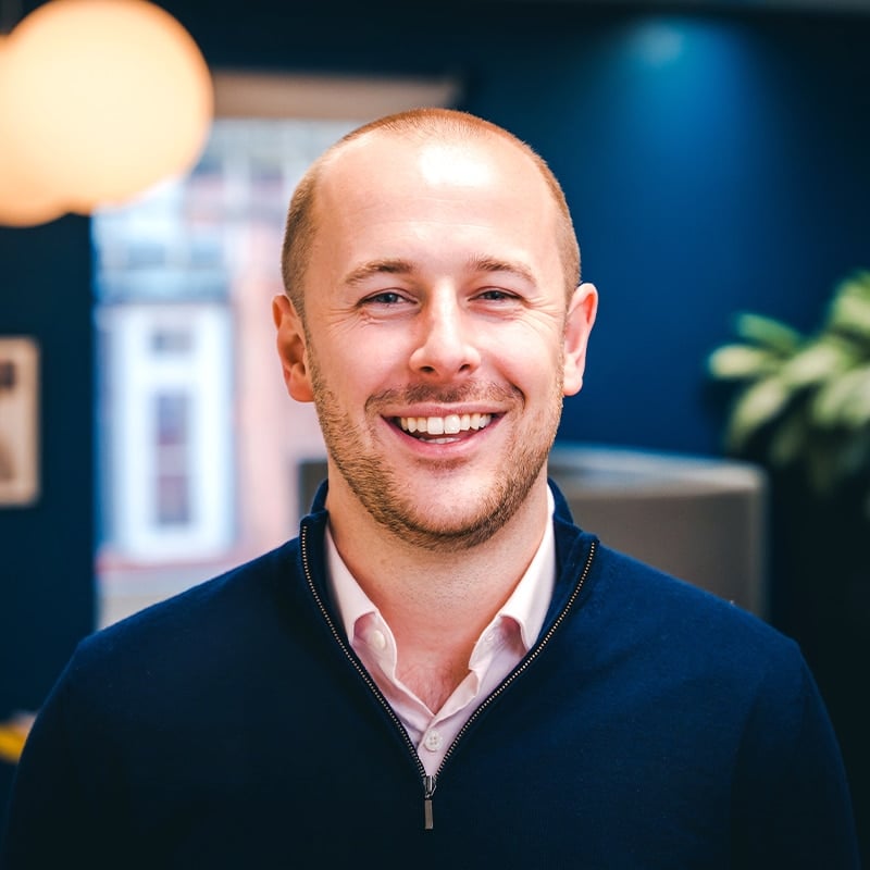 Headshot of Mathew Stent wearing a light pink shirt underneath a dark blue quarter-zip jumper in front of green plants and a dark blue wall.