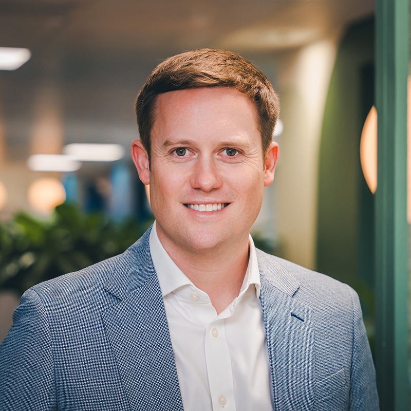 Headshot of Matt Whipp wearing a white shirt underneath a light blue blazer in front of a green wall and green plants.