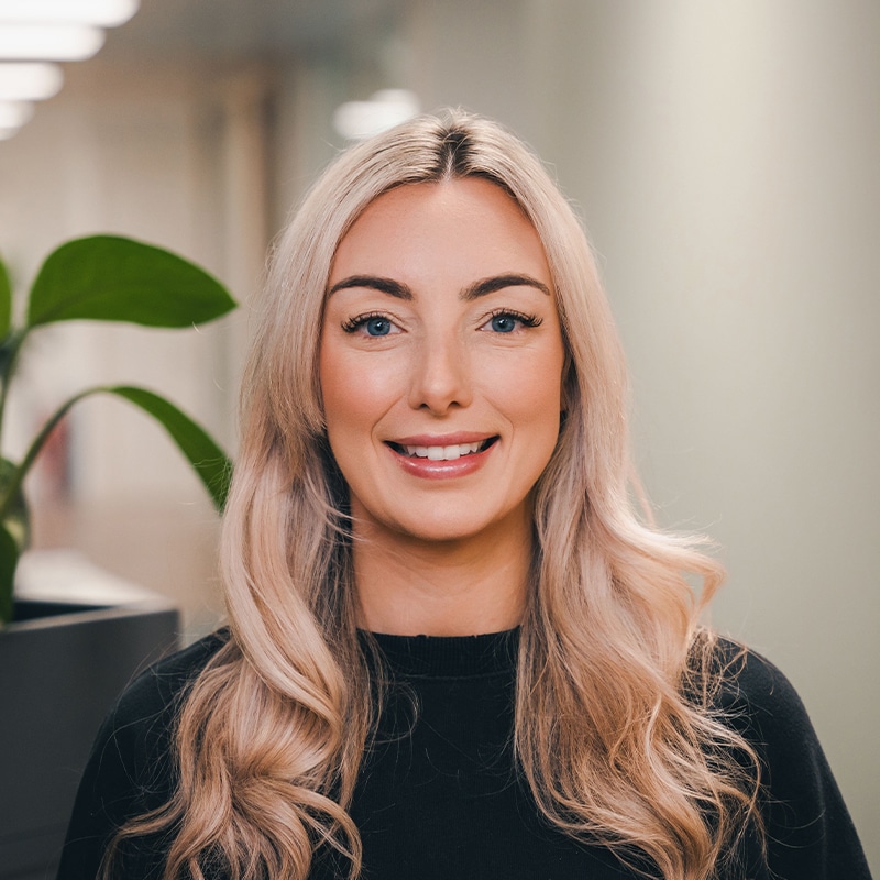 Headshot of Nicola Rogers wearing a black top with blonde curly hair in front of a white wall and green plants.