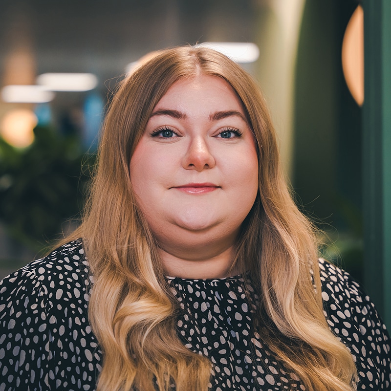 Headshot of Olivia Oakes-Dean, Finance Assistant at BIE, wearing a black and white spotty blouse in front of a green wall and green plants.