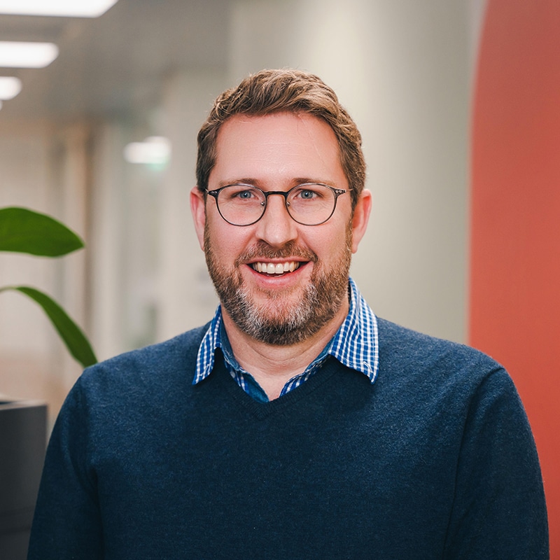 Headshot of Rob Knight wearing a blue and white checked shirt under a blue sweater in front of a pink and white wall and green plants.