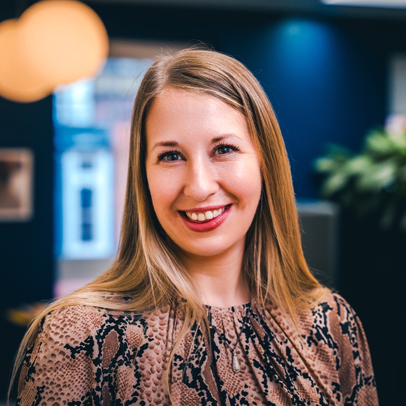 Headshot of Roz Alexander wearing a snake-print blouse in front of green plants and a blue wall.