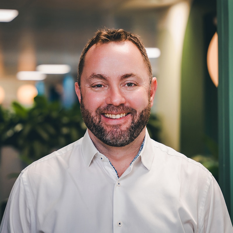 Headshot of Simon Moore wearing a white shirt in front of a green wall and green plants.