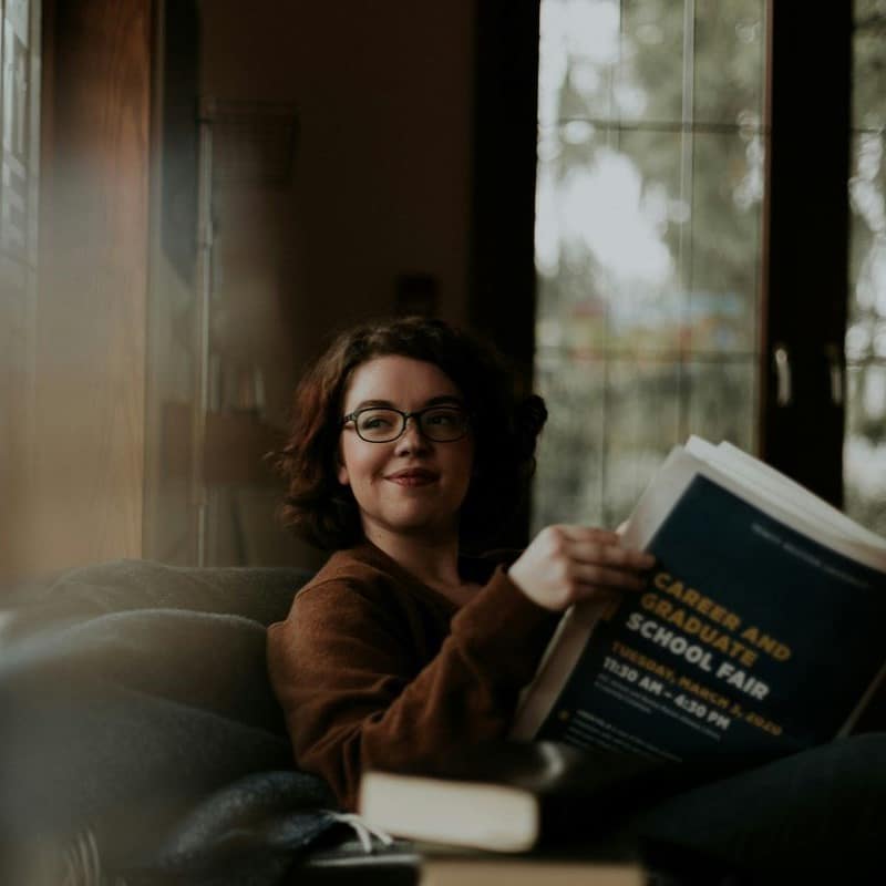 Woman sitting on a sofa reading a newspaper, wearing glasses and a long sleeved top.