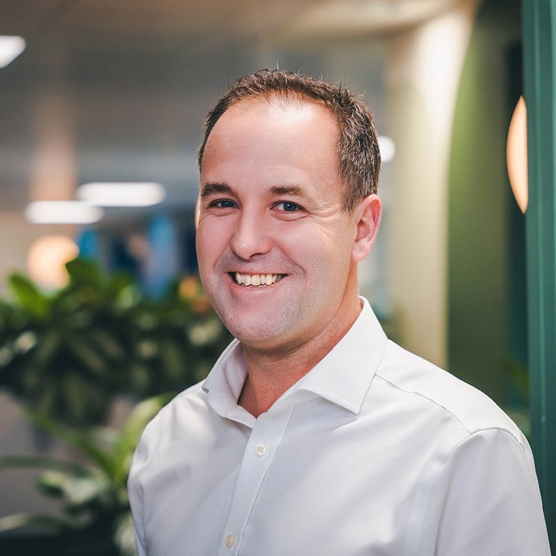 Headshot of Steve Arrow wearing a white shirt in front of a green wall and green plants.