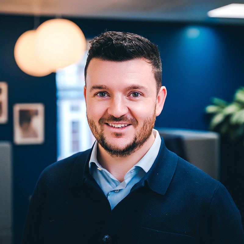 Headshot of Tom Hutchings, wearing a light blue shirt underneath a dark blue sweater in front of a grey seating area, green plants and a blue wall.