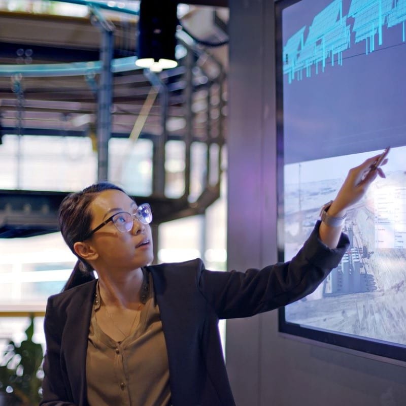 Young professional Asian woman wearing a black blazer and beige blouse. She is pointing at a screen of data.