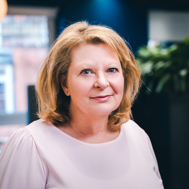 Headshot of Trish Alexander wearing a pink blouse with short blonde hair in front of a blue wall and green plants.