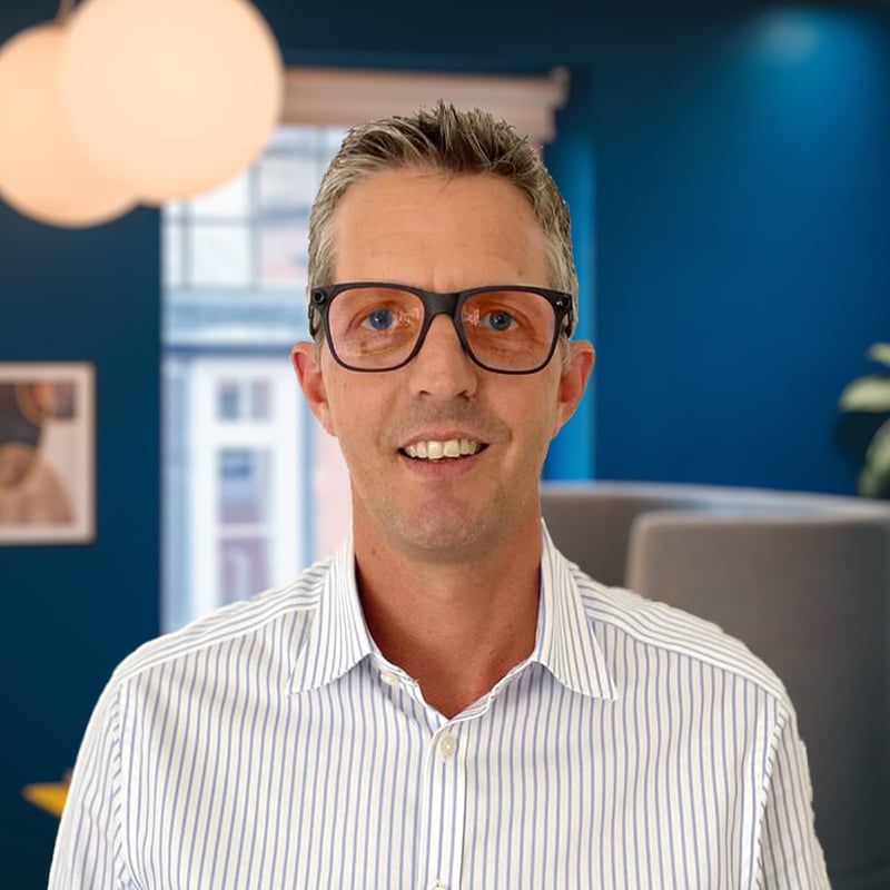 Headshot of Warren Pfotenhauer wearing a blue and white pinstripe shirt in front of a grey seat and blue walls.