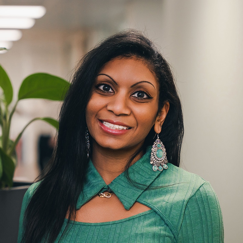 Headshot of Zoe Amenumey wearing a green top with tear-drop-shaped silver and green earrings in front of a white background and green plants.