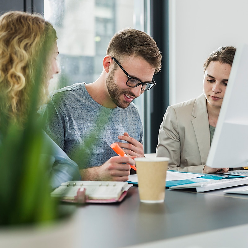 Three young colleagues using a highlighter on a document. Two are female and one is a male wearing glasses and a jumper.