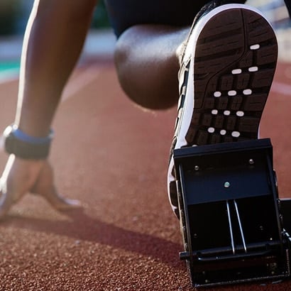 A black runner in the blocks ready to start a race. We can see the sole of their show and their hand poised on the left hand side.