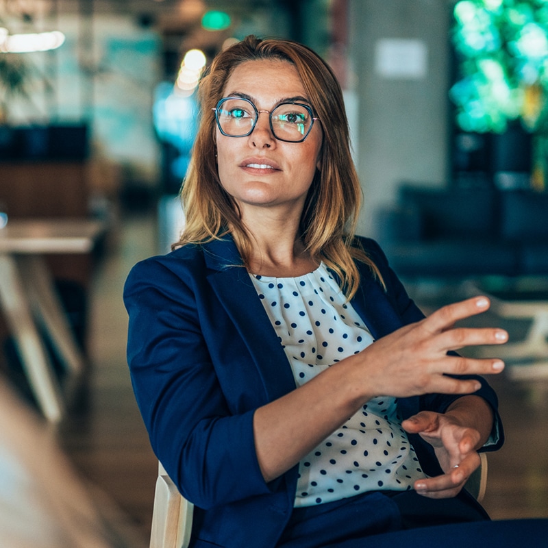 A young white female professional with blonde brown hair, wearing glasses and a polka dot top, with black spots on a white background, underneath a blue blazer. She appears to be in conversation with someone else out of shot.