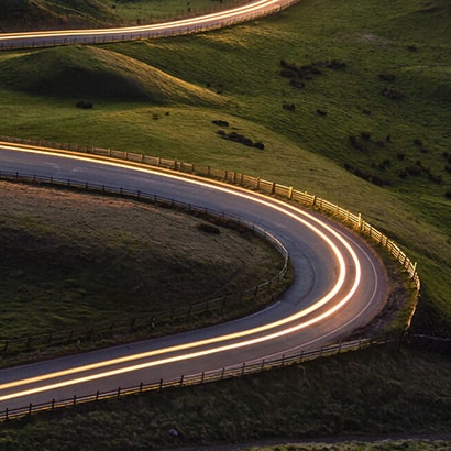 A winding road on green fields with a long exposure shot of two headlights travelling on the road.