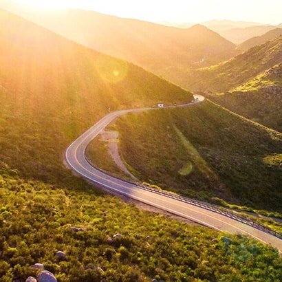 A winding road in green hills, with a sun flare coming from the top left hand corner.