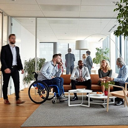 A group of colleagues of different ethnicities in a modern office sat on a sofa and chairs in a semi-circle talking. One male employee is in a wheelchair.