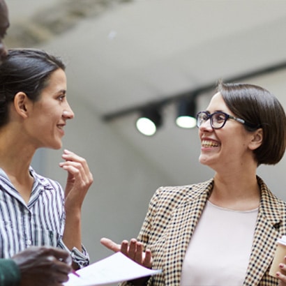 Two business women having a conversation and smiling, Both are middle aged with dark hair. One is wearing glasses and a checked blazer, the other is in a stripey blue and white shirt.